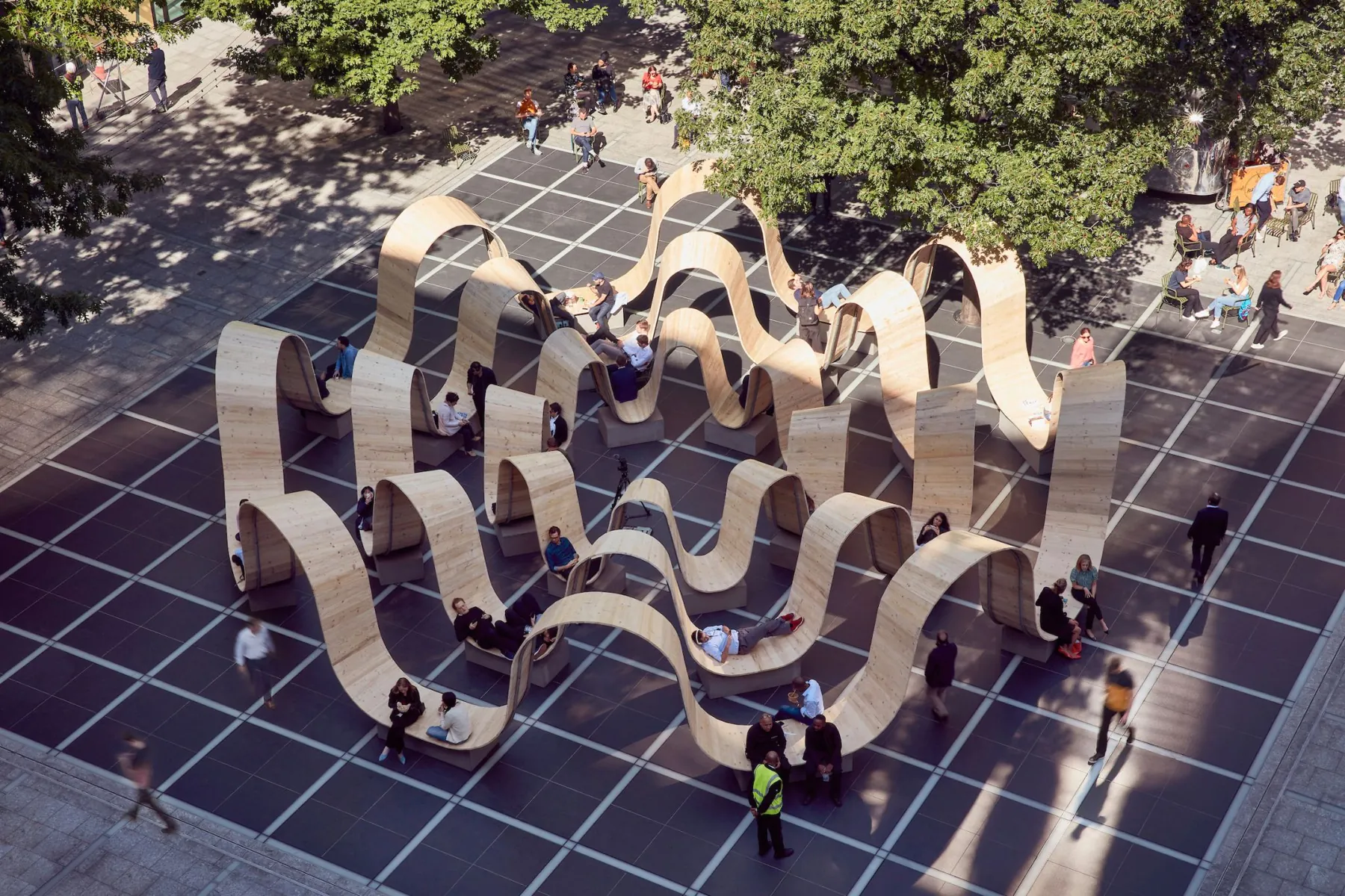 Aerial view of Please Be Seated in London: undulating timber ribbons that people sit, lie and walk on across a paved square.