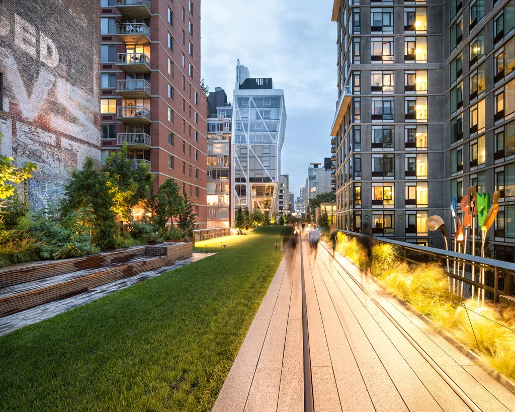 The High Line in New York at dusk: a planted walkway with timber benches between apartment buildings, lit by warm low-level lighting.