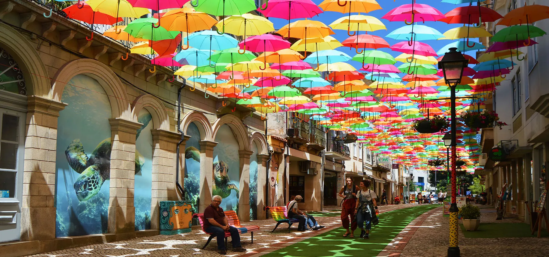 Umbrella Sky Project in Águeda, Portugal: a canopy of colourful umbrellas suspended over a pedestrian street, with people sitting on painted benches below.