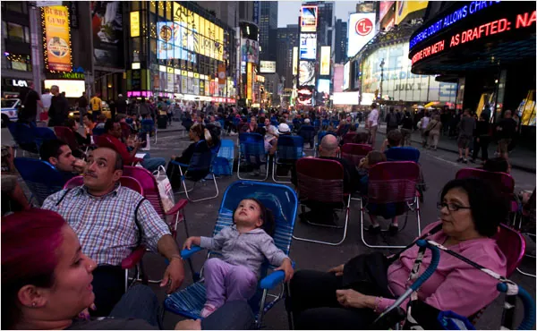 People relaxing in folding lawn chairs on a car-free Times Square at night, surrounded by lit billboards.