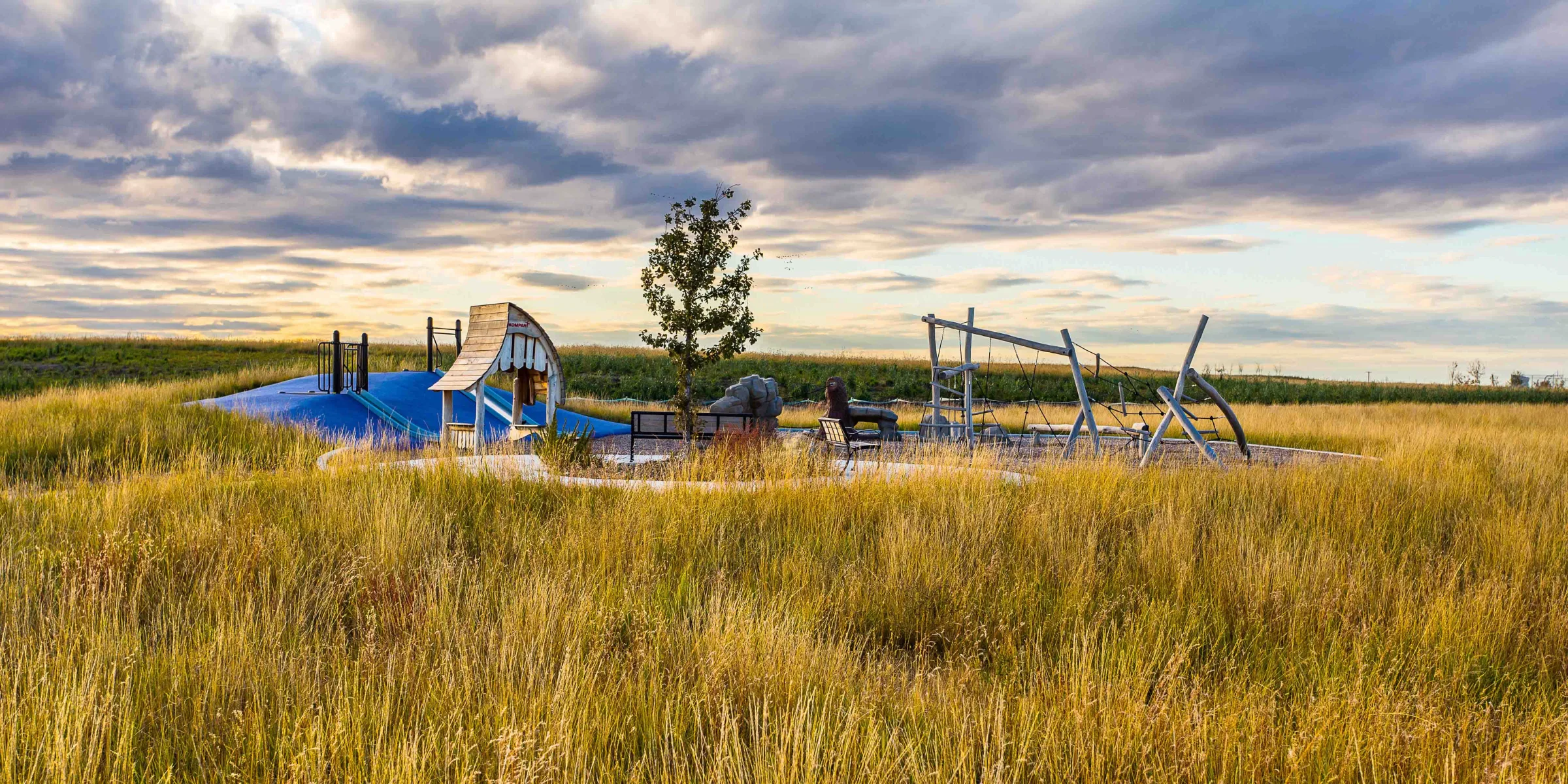 Shaheed Bhagat Singh Nature Playground: a blue slide mound, timber lookout and rope climbing frame set in tall prairie grass under a wide evening sky.