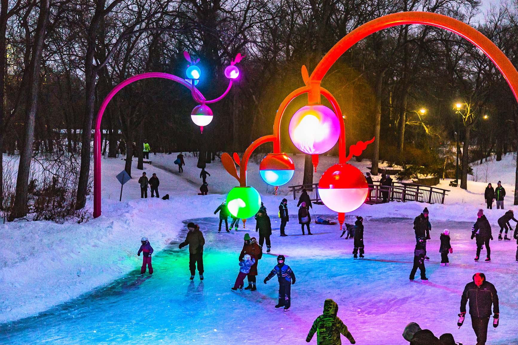 Bokeh at Kildonan Park: red sculptural arches holding glowing coloured orbs over a skating trail, with skaters on the ice at dusk.