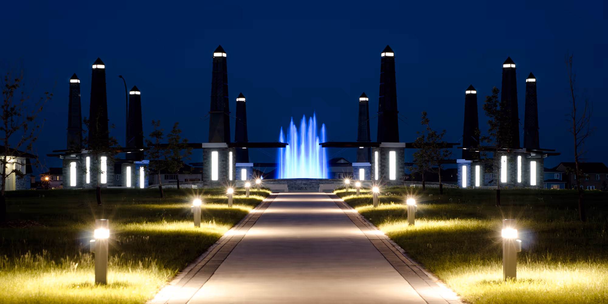 Water Bend Park fountain at night in Winnipeg: a blue-lit water jet framed by two rows of tall obelisk light columns along a paved promenade.